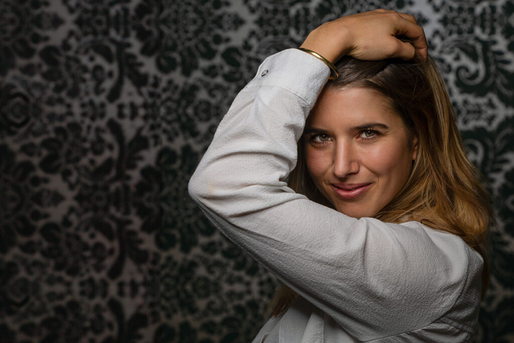Eine junge Frau mit langen Haaren posiert selbstbewusst und trägt ein weißes Hemd. Sie hat den Arm über den Kopf erhoben und lächelt leicht vor einem gemusterten dunklen Hintergrund.
