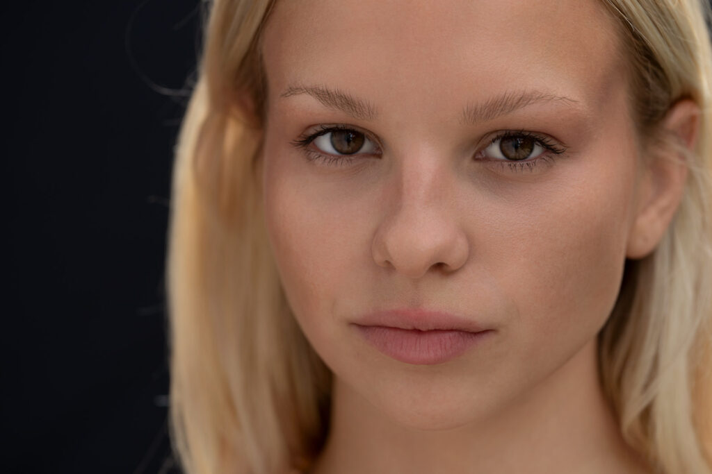 Ein Nahaufnahme-Portrait einer jungen Frau mit langen blonden Haaren vor einem dunklen Hintergrund. Die Person hat einen neutralen Gesichtsausdruck, ist dezent geschminkt und hat braune Augen. Sie blickt direkt in die Kamera.